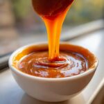 Close-up of thick, glossy caramel sauce being poured from a spoon into a small white bowl.