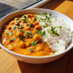 A white bowl filled with vibrant orange pumpkin curry featuring chickpeas, served next to fluffy white rice and garnished with cilantro.