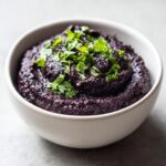 A close-up of thick, dark purple black bean dip served in a light bowl and garnished with fresh cilantro.
