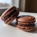 Two dark brown chocolate macarons filled with rich ganache, one sprinkled with sea salt.