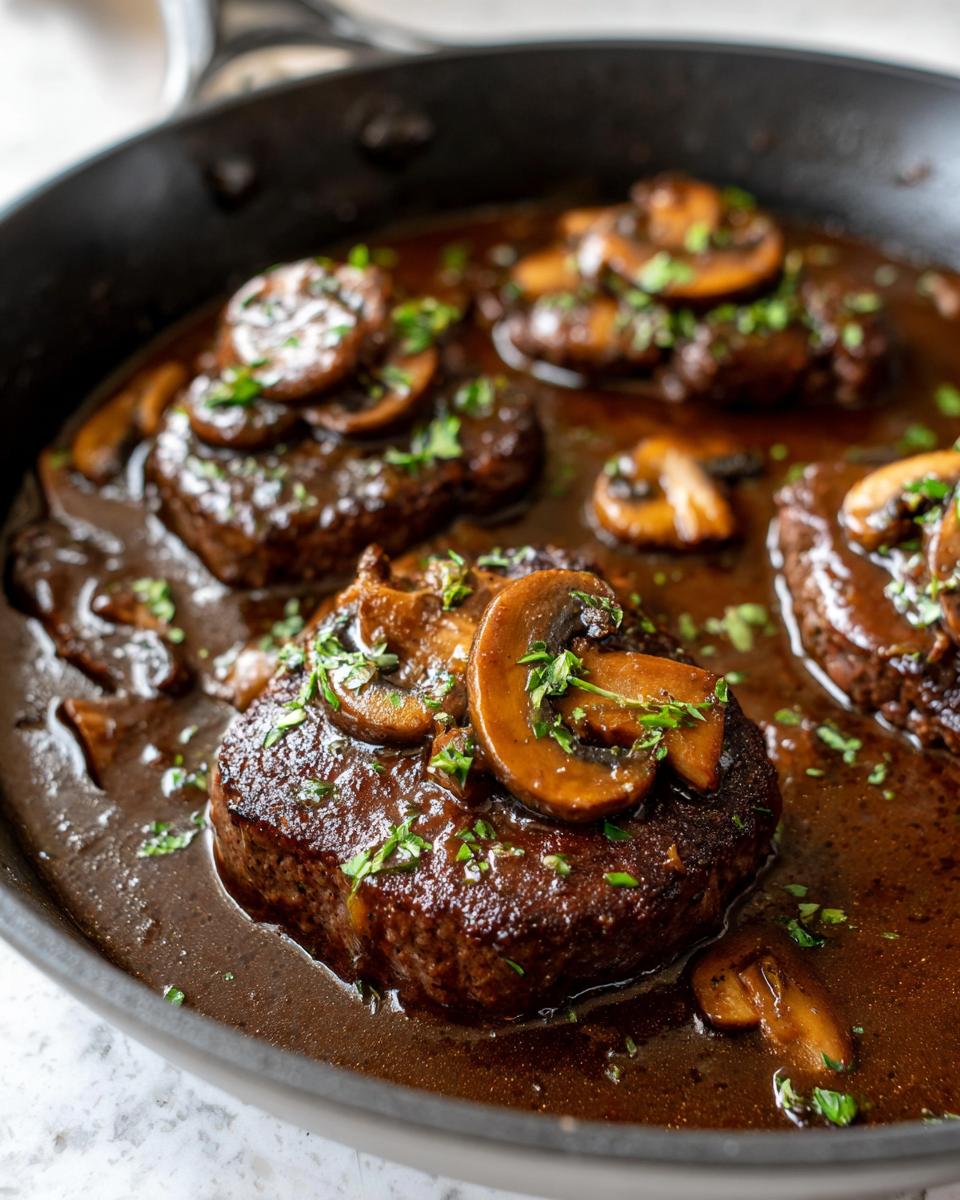 Close-up of juicy Salisbury steak patties covered in mushroom gravy and fresh parsley in a black skillet.