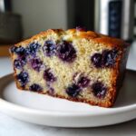 A thick slice of moist blueberry banana bread showing bright purple blueberries baked into the golden crumb.