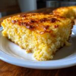 Close-up of a golden slice of baked cheese grits with a caramelized top on a white plate.