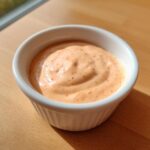 A small white ramekin filled with creamy, orange-pink smash burger sauce speckled with spices.