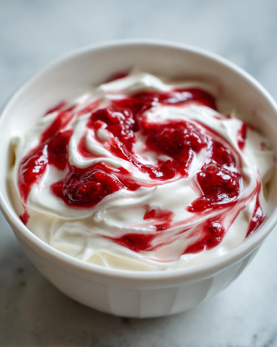 Close-up of creamy white strawberry cheesecake dip swirled with bright red strawberry topping in a white bowl.