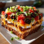 A square slice of the ultimate 7 layer dip showing distinct layers topped with cheese, tomatoes, olives, and green onions.