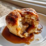 A warm apple dumpling broken open to show the spiced apple filling, drizzled generously with rich caramel sauce.