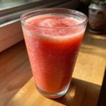 A tall glass filled with a vibrant red, icy watermelon smoothie sitting on a wooden surface near a window.