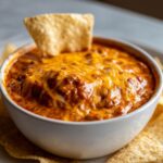 Close-up of a white bowl filled with hot, bubbly chili cheese dip topped with melted cheddar cheese, served with a tortilla chip.