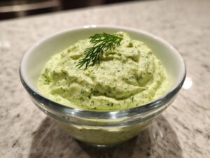 A close-up of creamy, green dill dip topped with a fresh sprig of dill, served in a clear glass bowl.