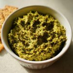 Close-up of thick, vibrant green olive dip served in a white bowl next to crackers.