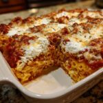 Close-up of a rich, cheesy baked ziti pasta bake with a portion removed, showing layers of sauce and ricotta.
