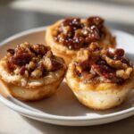 Three golden-crusted pecan tassies filled with gooey, caramelized pecans sitting on a white plate.