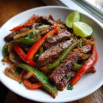 Close-up of sizzling steak fajitas with sliced steak, red and green peppers, and onions, garnished with lime wedges.