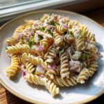 A close-up of creamy Tuna Pasta Salad mixed with fusilli pasta, red onion, and celery on a light plate.
