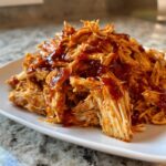 A mound of tender, shredded pulled chicken generously drizzled with rich barbecue sauce on a white plate.