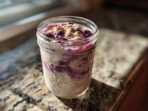 A jar of creamy blueberry overnight oats topped with fruit and nut butter, sitting on a sunny granite countertop.