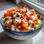 A close-up of a buffalo chicken bowl featuring orange buffalo-coated chicken pieces drizzled with white ranch dressing over rice and lettuce.