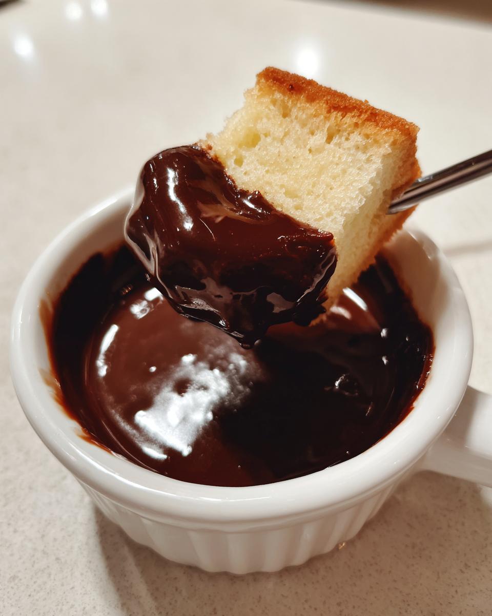 A piece of sponge cake being dipped into rich, melted chocolate fondue in a white ramekin.