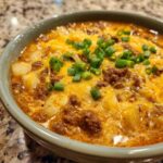 Close-up of a steaming bowl of cheeseburger soup recipe, loaded with ground beef, potatoes, melted cheddar cheese, and green onions.