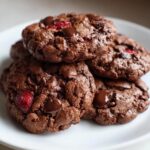 A close-up stack of rich, dark chocolate cherry cookies dotted with melted chocolate chips and bright red cherries.