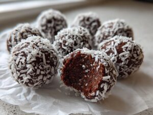 A close-up of several rich, dark chocolate coconut balls rolled in shredded coconut, one with a bite taken out.