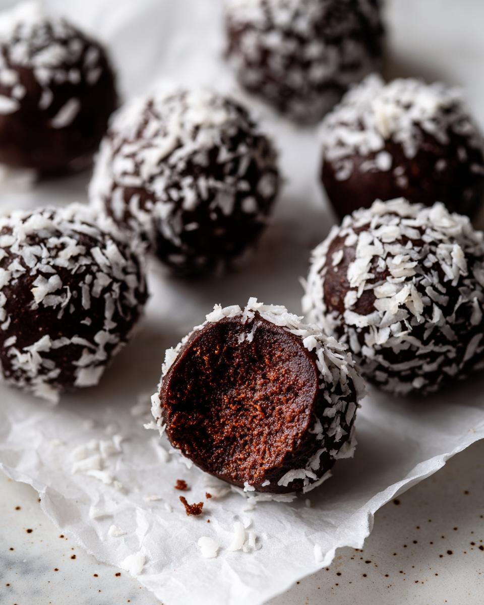 Close-up of rich chocolate coconut balls rolled in shredded coconut, one is bitten to show the dense interior.