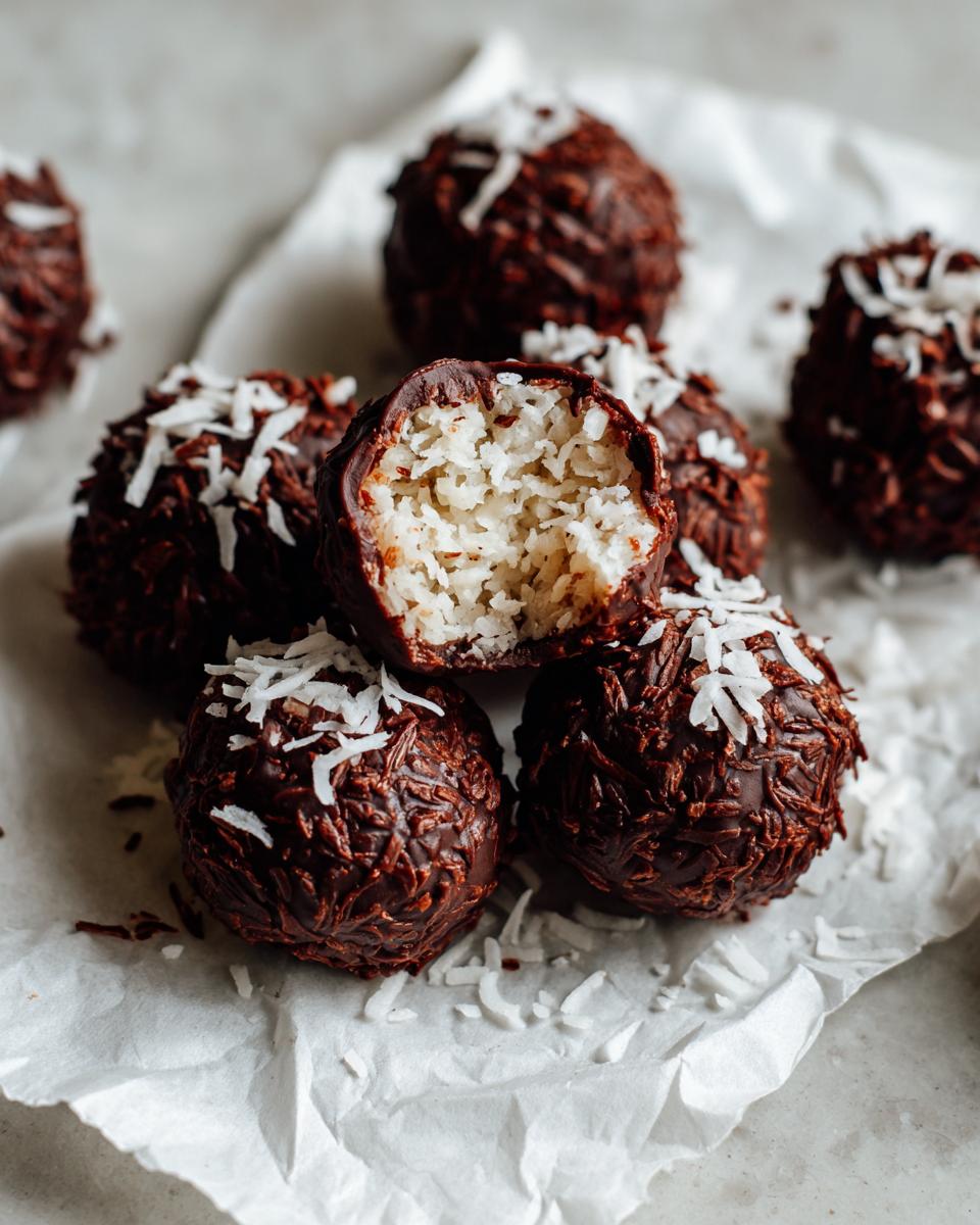 A pile of rich chocolate covered coconut balls, one is bitten open revealing the white shredded coconut filling.