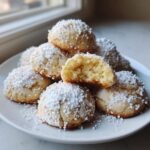 A stack of soft Christmas gooey butter cookies covered in white sanding sugar, one cookie is broken open showing the chewy center.