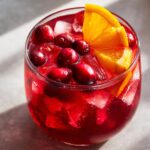 Close-up of a glass filled with bright red thanksgiving punch, ice, fresh cranberries, and an orange slice.