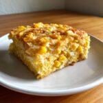 A square serving of moist cream cheese corn casserole with visible corn kernels, served on a white plate.