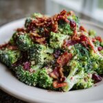 A close-up of creamy broccoli salad topped with crispy bacon pieces and dried cranberries.