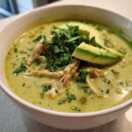 A close-up of a bowl of creamy green chicken poblano soup topped with shredded chicken, avocado slices, and cilantro.