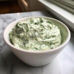 A close-up of a white bowl filled with creamy, light green dill dip generously topped with fresh dill.