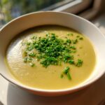 A close-up of creamy potato leek soup in a white bowl, generously garnished with bright green chopped chives.