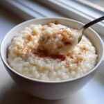 A spoonful of creamy rice pudding topped with cinnamon being lifted from a white bowl of rice pudding.