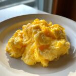 Close-up of fluffy, bright yellow scrambled eggs served on a white plate, highlighted by natural sunlight.