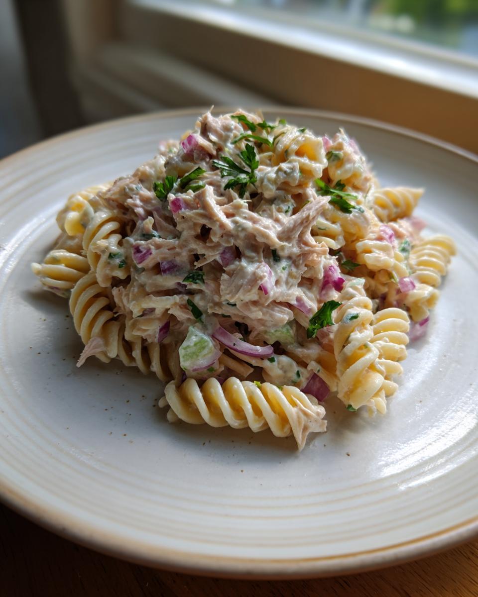 A close-up serving of creamy Tuna Pasta Salad featuring rotini pasta, flaked tuna, and diced red onion, garnished with parsley.