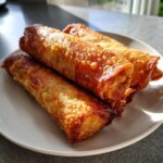Close-up of four golden brown, crispy fried egg roll appetizers stacked on a white plate.