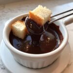 A piece of pound cake is dipped into rich, melted chocolate fondue using a fondue fork.