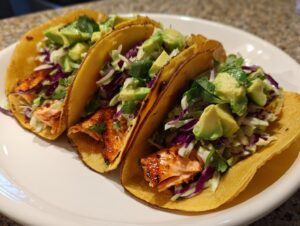 Three fully loaded salmon tacos featuring flaky seasoned salmon, purple cabbage slaw, and diced avocado on corn tortillas.