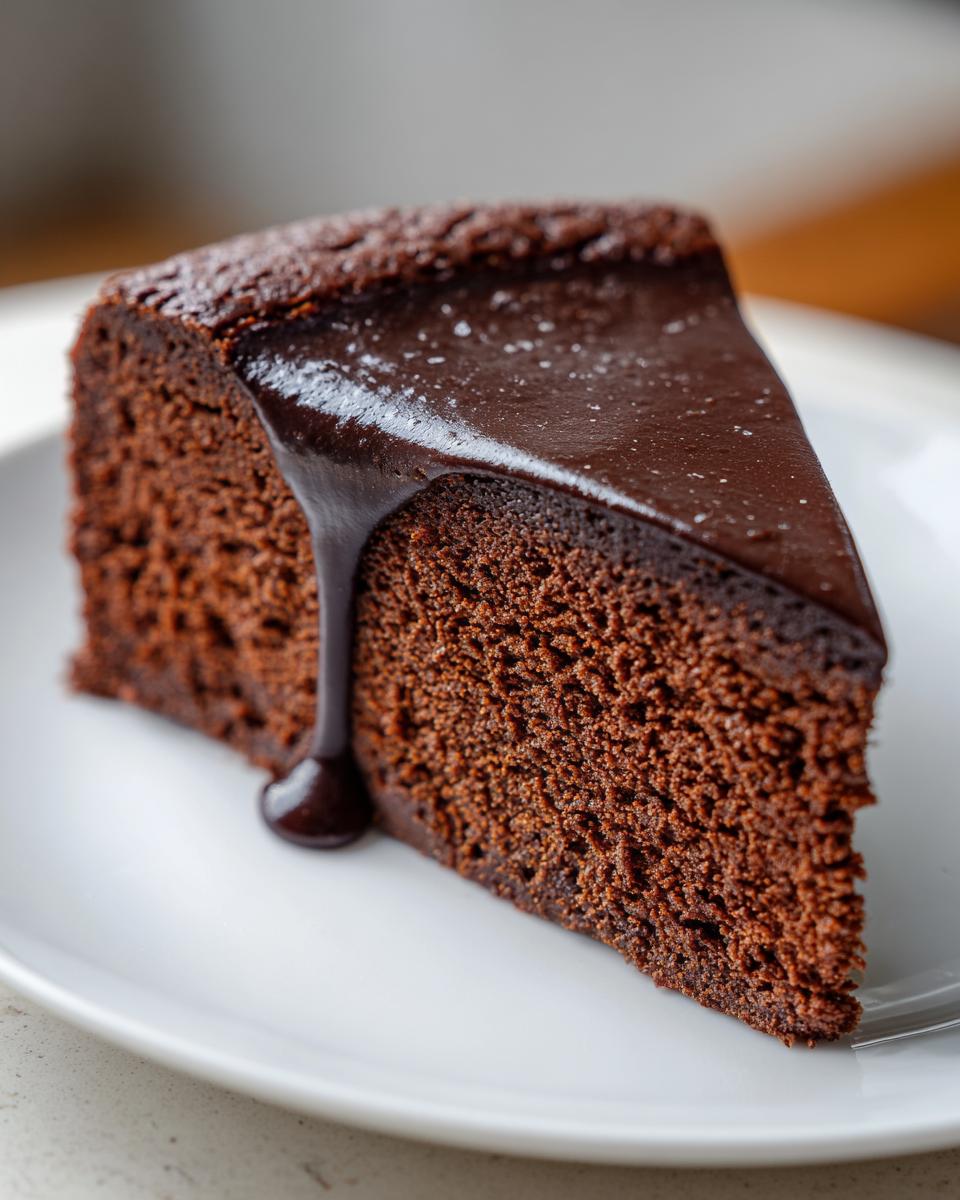 A close-up of a dense slice of flourless chocolate cake topped with dripping dark chocolate ganache.