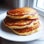 A close-up stack of three golden-brown, fluffy banana oatmeal pancakes on a white plate.