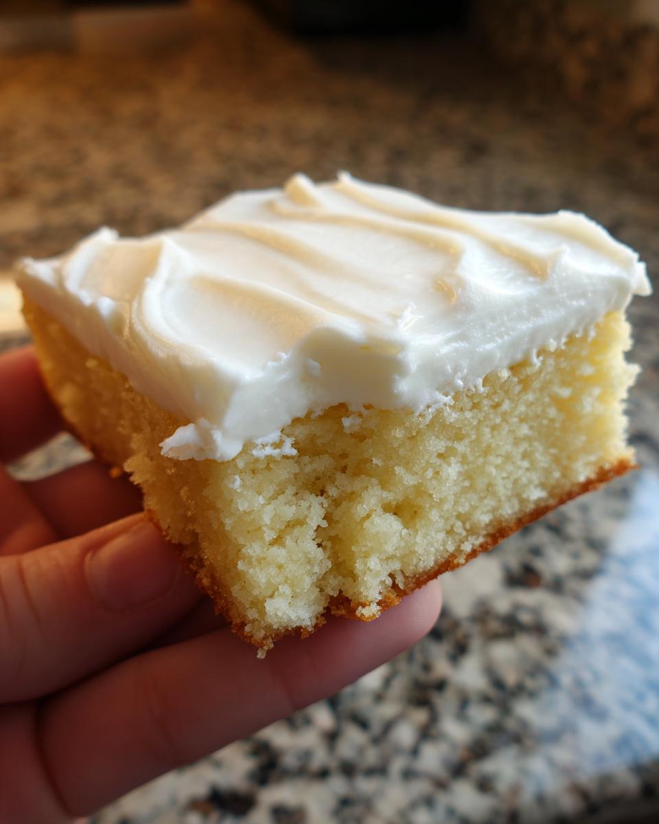 A hand holding a square piece of soft, thick sugar cookie bars topped with thick white frosting.