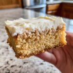 A close-up view of a thick, moist slice of sugar cookie bars topped with creamy white frosting, held by a hand.