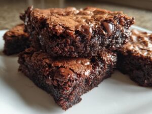 Close-up of several fudgy brownies stacked on a white plate, showing melted chocolate chips and a shiny top crust.