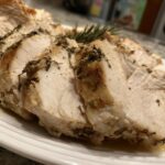 Close-up of thick, juicy slices of herb-seasoned crockpot turkey breast resting on a white plate.