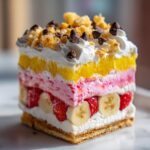 A tall, square slice of banana split cake showing distinct layers of crust, bananas, strawberries, pink mousse, yellow topping, and whipped cream with chocolate chips.