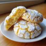 Three bright yellow lemon crinkle cookies heavily dusted with powdered sugar stacked on a white plate.