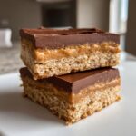 Two stacked squares of lunch lady peanut butter bars featuring a crust, peanut butter layer, and chocolate topping.